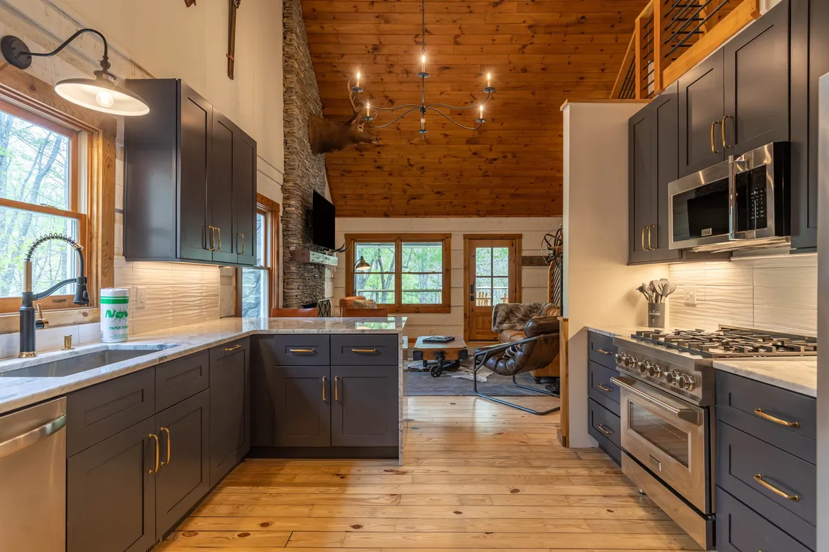 An open-concept area showcasing dark cabinetry, stainless steel appliances, a cozy stone fireplace, and large windows letting in natural light. The space transitions seamlessly between kitchen and living, blending rustic charm with contemporary finishes.