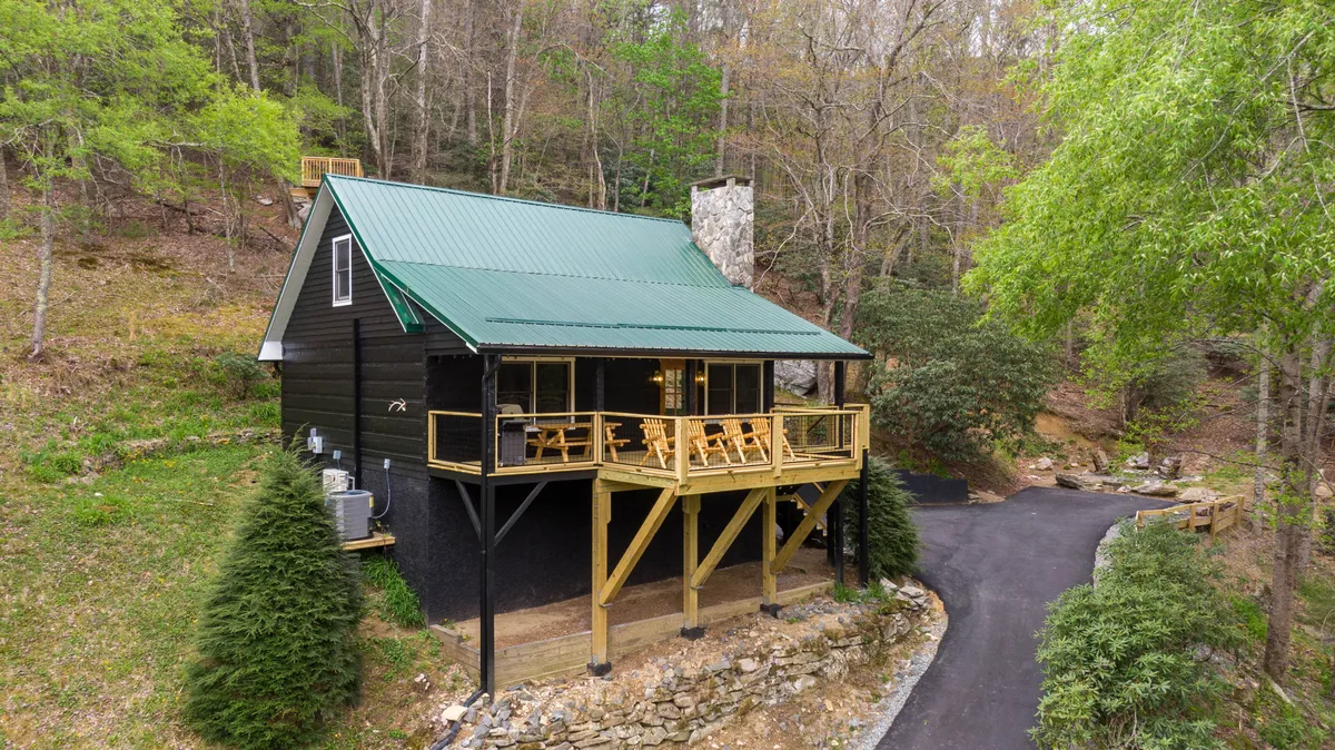 This exterior shot captures a welcoming log cabin with a spacious wooden deck overlooking the lush banks of the Watauga River. The green metal roof and striking stone chimney stand out amidst surrounding trees, while the golden glow of sunset sets a tranquil mood perfect for relaxation and gatherings.