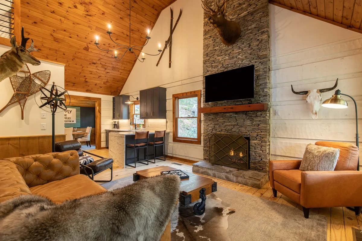 A spacious living area featuring a striking stone fireplace, rustic wooden beams, and plush seating, perfect for socializing or relaxing after a day of adventure. A large chandelier adds warmth to the space, and a mounted deer head above the fireplace provides a touch of mountain lodge charm.