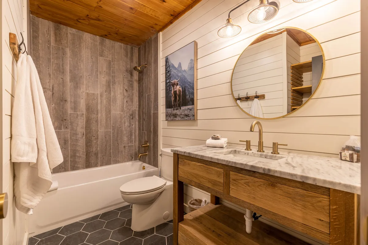 A spa-inspired bathroom with a wooden vanity, round mirror, striking artwork of a cow, and textured hexagonal floor tiles. The space exudes calm and rustic charm, inviting guests to enjoy relaxing moments and modern amenities.