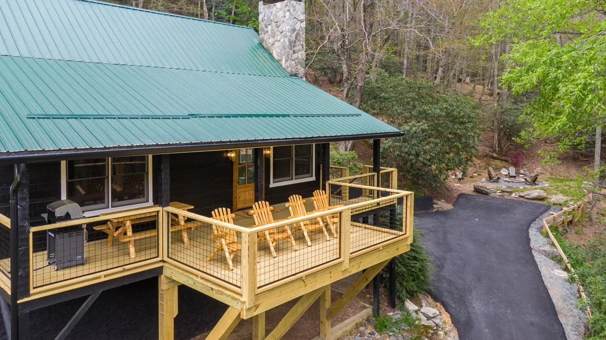 A spacious wooden deck with Adirondack chairs and a view of the surrounding nature, featuring a stone chimney and a green roof. The arrangement invites guests to relax, sip coffee, and take in the tranquil mountain atmosphere.
