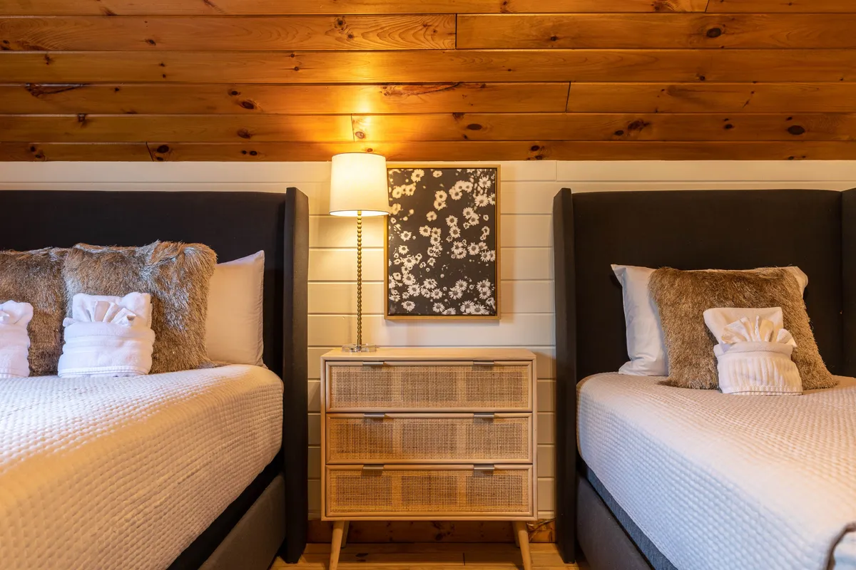 A cozy bedroom with two beds, a nightstand and lamp, and framed artwork. The setup is perfect for friends or family sharing a space, blending comfort with a welcoming, relaxed atmosphere.