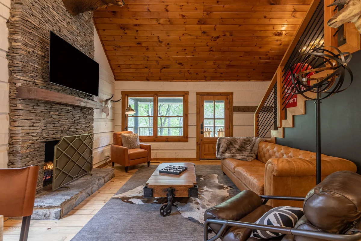 A spacious great room featuring a dramatic stacked stone fireplace as its centerpiece, flanked by plush leather sofas. Large windows bathe the space in natural light, and the wood-paneled ceiling soars overhead, showcasing the log cabin's craftsmanship. A cowhide rug and tasteful decor create a warm, inviting ambiance.