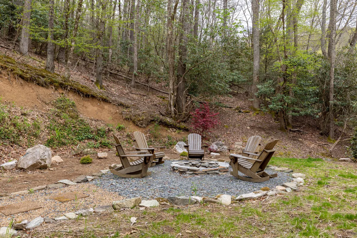 An inviting fire pit area is surrounded by Adirondack chairs, set against a backdrop of lush trees and a gravel pathway. The scene evokes cozy evenings by the fire, storytelling, and connecting under the stars.