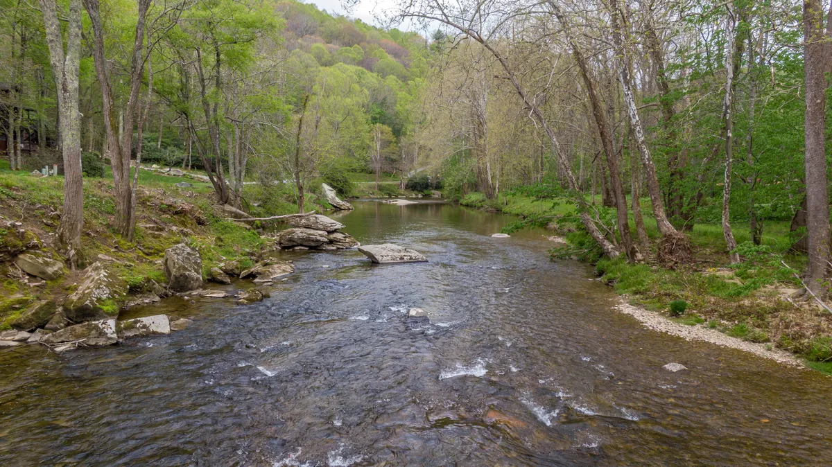 A serene, tree-lined creek with rock formations meanders through the landscape, inviting guests to fish, hike, or simply enjoy the peaceful rhythms of nature. The surrounding trees suggest a vibrant natural habitat and the gentle flow of water hints at the adventure awaiting just steps from the cabin.