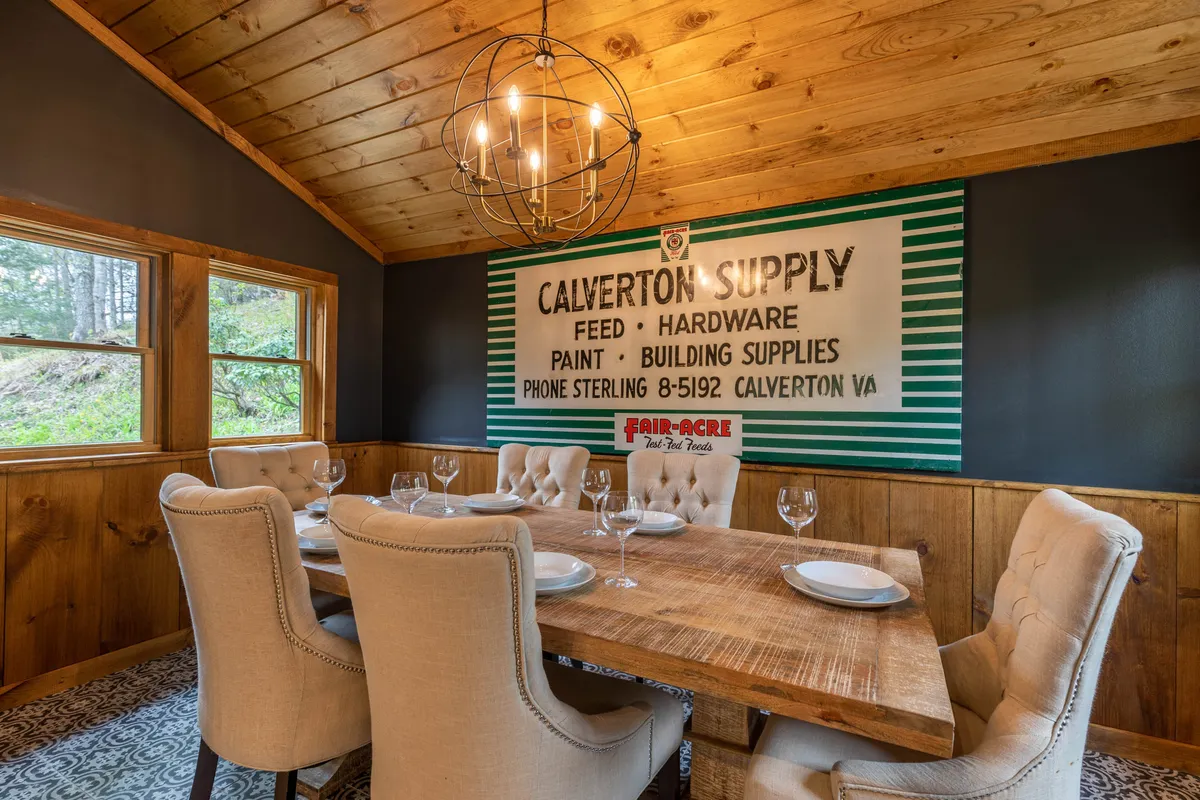 A welcoming dining area with a solid wooden table beneath a stylish chandelier, accented by a vintage 'Calverton Supply' sign on the wall. The space radiates warmth and hospitality, ideal for shared meals or friendly game nights.