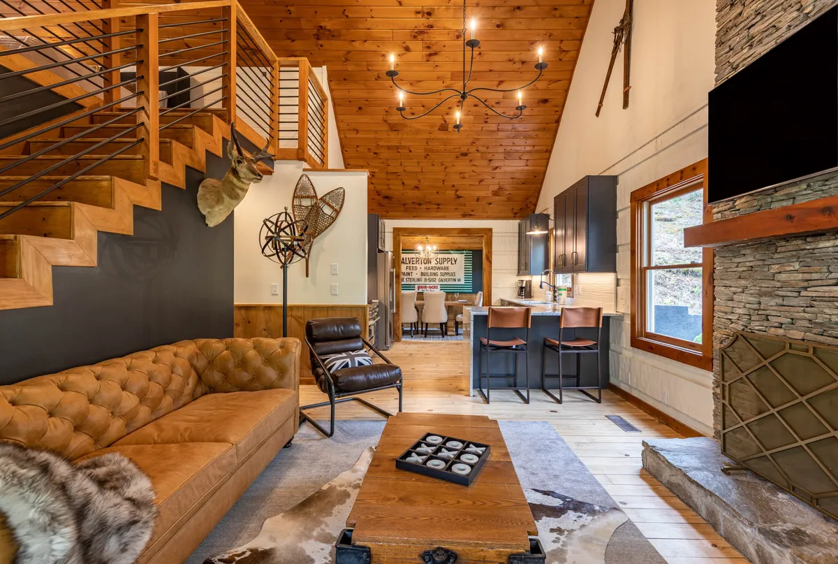A warm and spacious living area featuring a striking stone fireplace, a plush leather sofa, a rustic wooden staircase, and decorative wall art. The centerpiece chandelier casts a cozy glow, while the seating invites relaxation, conversation, or family movie nights.