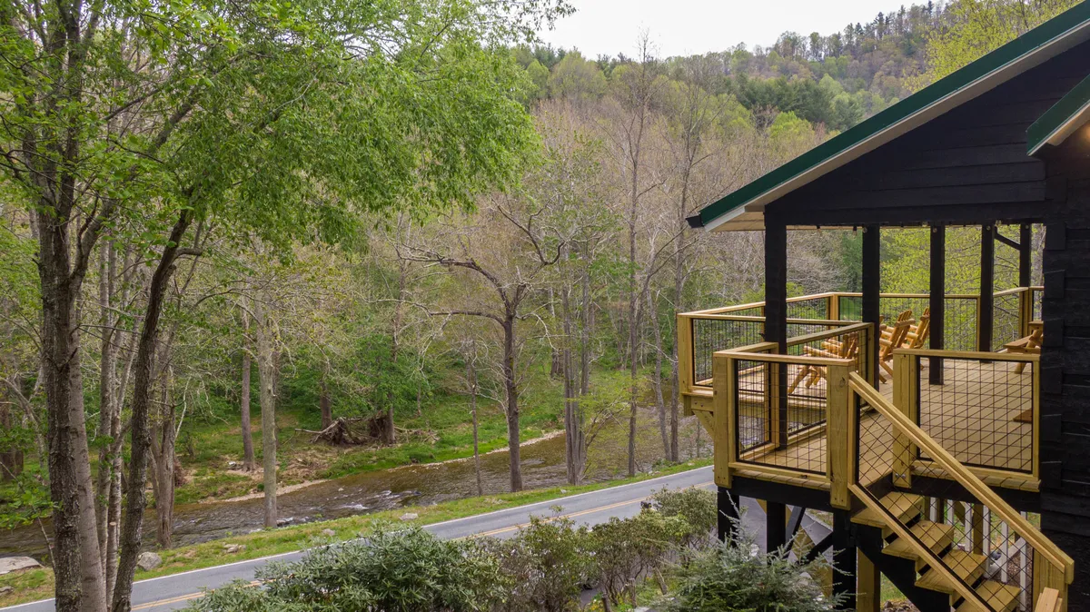 This image presents the cabin’s deck with a wooden railing overlooking a tranquil stream and surrounded by lush trees. The setting conveys a peaceful morning or evening atmosphere, ideal for relaxing while immersed in nature.