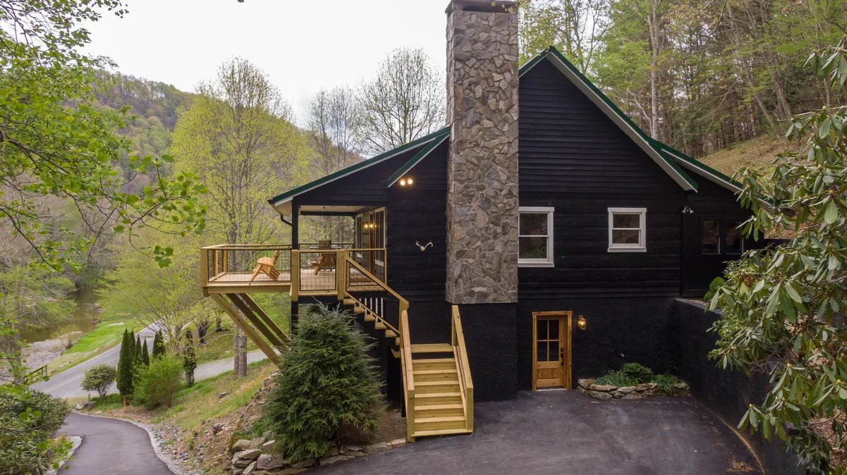 This exterior shot captures the cabin’s stone chimney, expansive wooden deck, front entrance, and the surrounding greenery, set against the soft glow of late afternoon. The inviting outdoor space hints at both daytime relaxation and the warmth of evening gatherings.