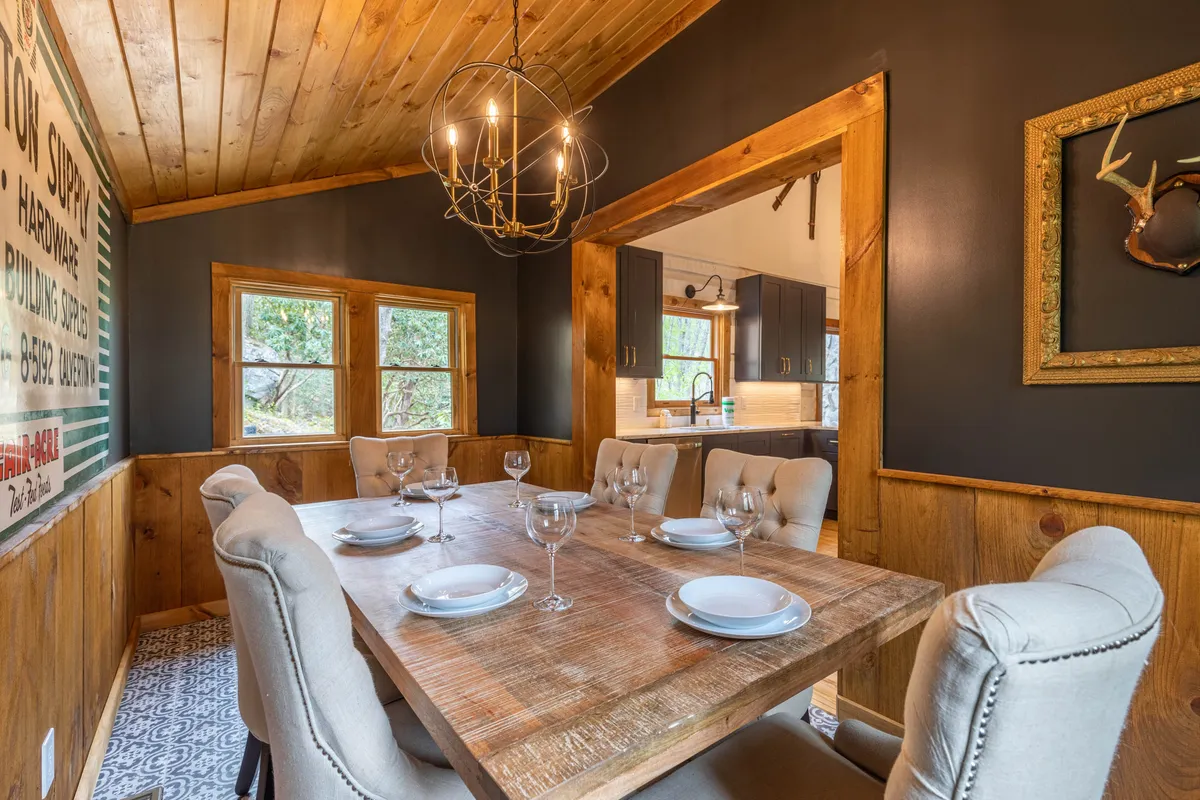 A warmly lit dining area with a rustic wooden table, elegant chandelier, and large windows inviting natural light. The decor includes tasteful wall art, creating a welcoming environment that encourages shared moments and conversation.