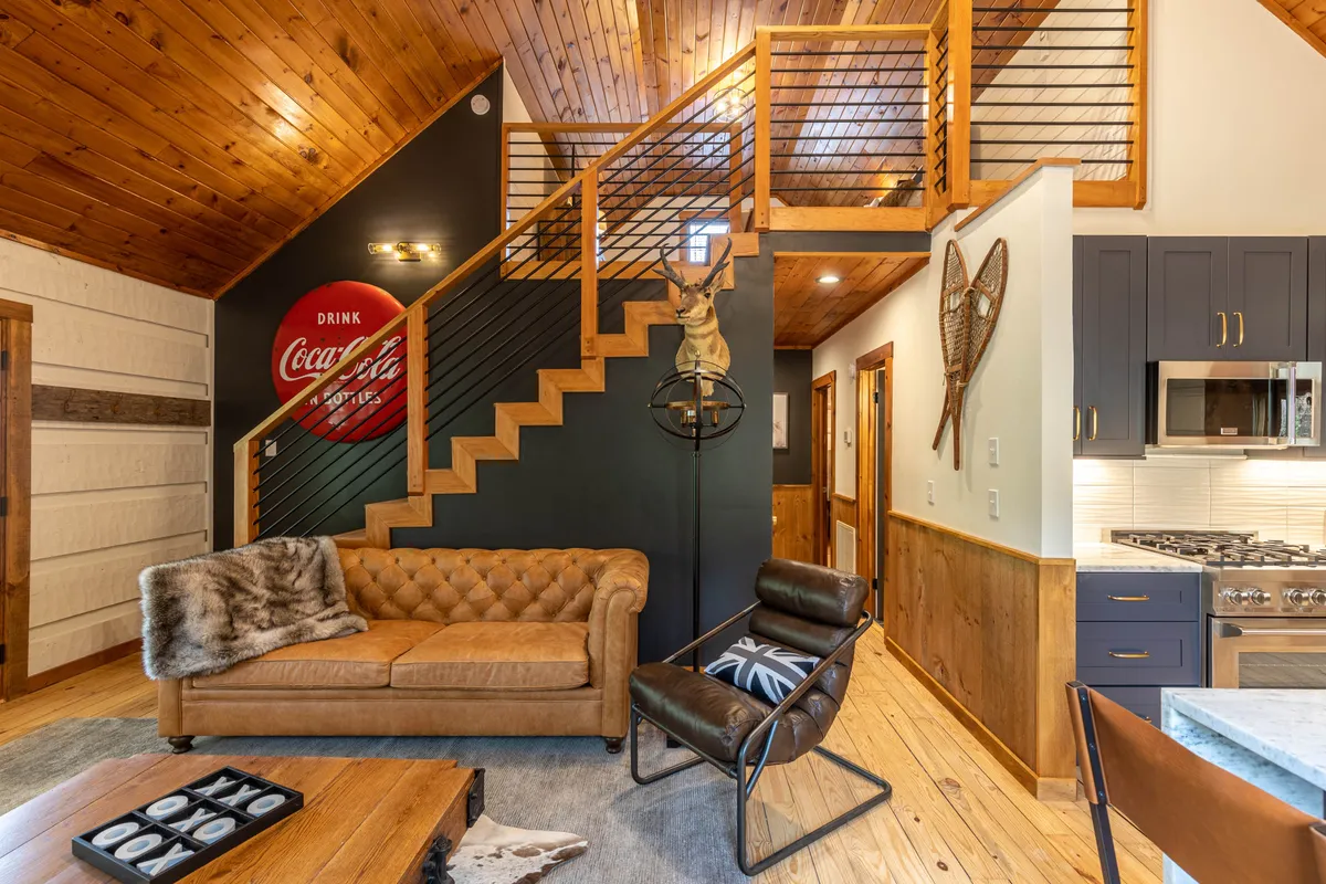A cozy angle into the great room and kitchen, featuring a Chesterfield sofa positioned near the staircase, a Coca-Cola sign adding a playful touch, and a clear view into the modern kitchen and dining area. The scene feels lived-in and stylish, emphasizing both relaxation and the social heart of the home.