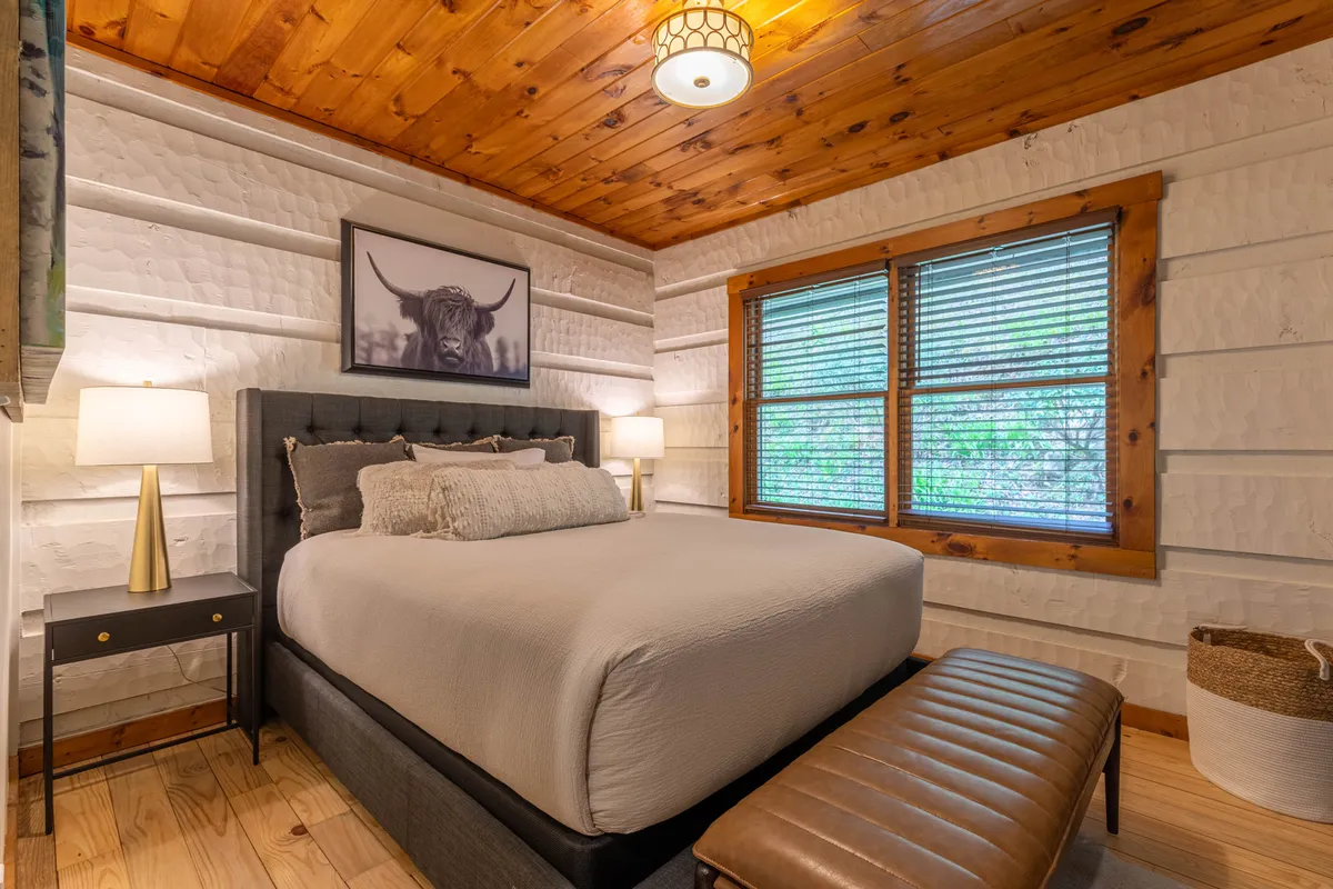 A tranquil bedroom with a queen-sized bed is softly lit by natural light streaming through a window. The space features elegant nightstand lamps, calming artwork of a bull above the bed, and plush bedding, all framed by rustic wooden accents that emphasize comfort and warmth.